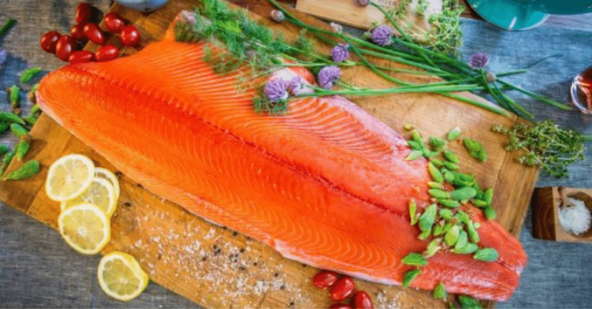 Large raw salmon fillet on wood cutting board with herbs, cherry tomatoes and lemon slices scattered around