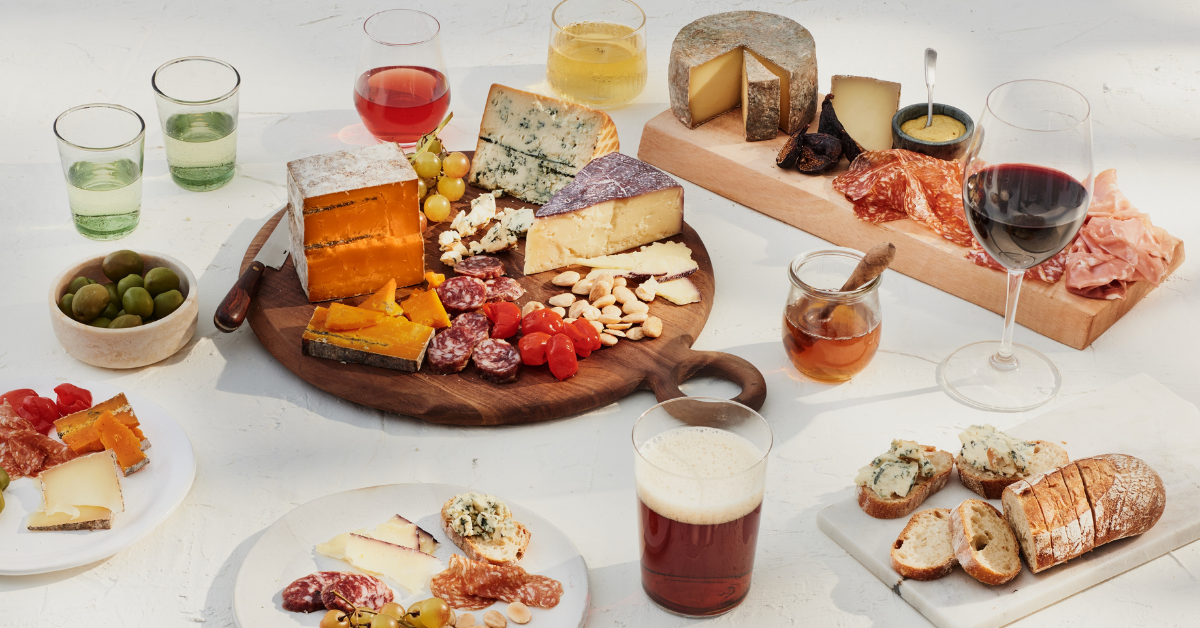 Wood serving boards and white plates with variety of cheeses, meats and fruits, with glasses of wine and beer arranged around them