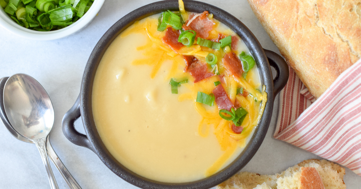 Creamy potato soup in small black crock topped with shredded cheese, bacon pieces, and sliced green onions