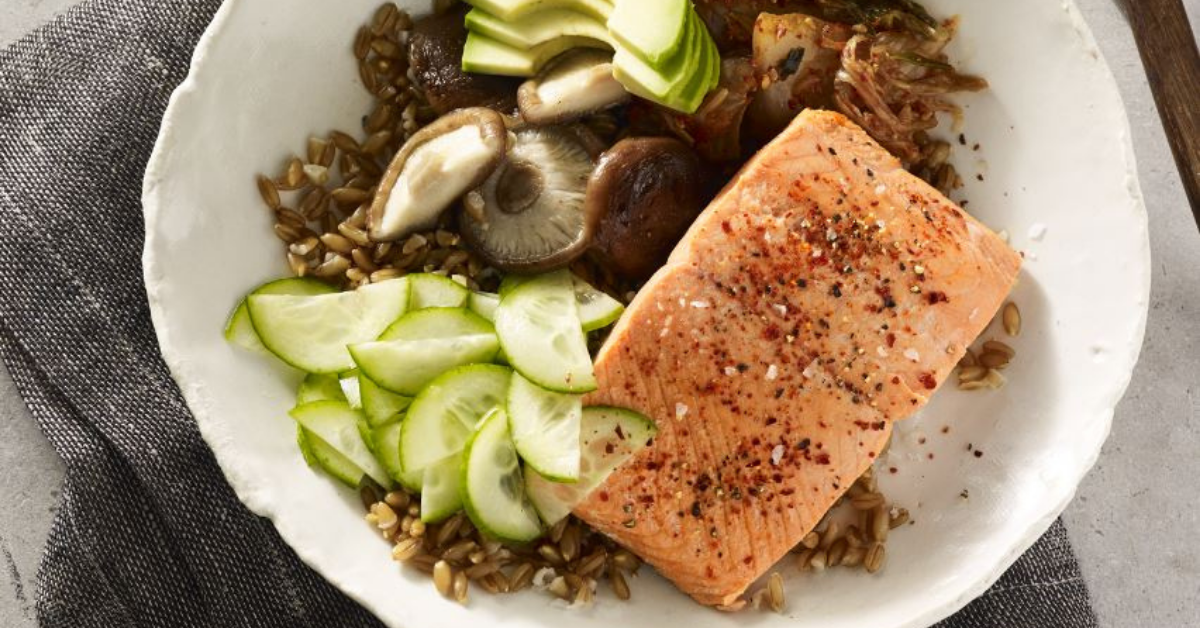 Salmon fillet, sliced cucumber, sliced avocado and mushrooms on bed of grains in white bowl