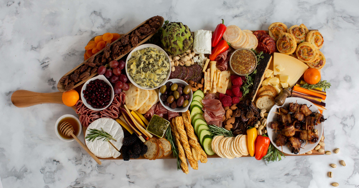 Wood serving board filled with various cheese, nuts, fruits, vegetables, dips, and crackers