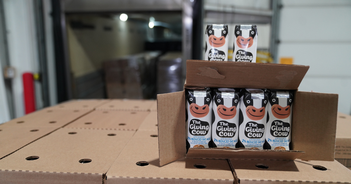 Open cardboard box of The Giving Box milk cartons on top of stack of boxes in a warehouse
