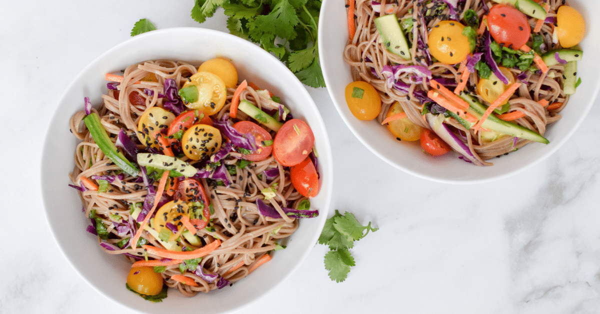 Noodles topped with halved tomatoes, sliced cucumbers, shredded cabbage and black sesame seeds in white bowls